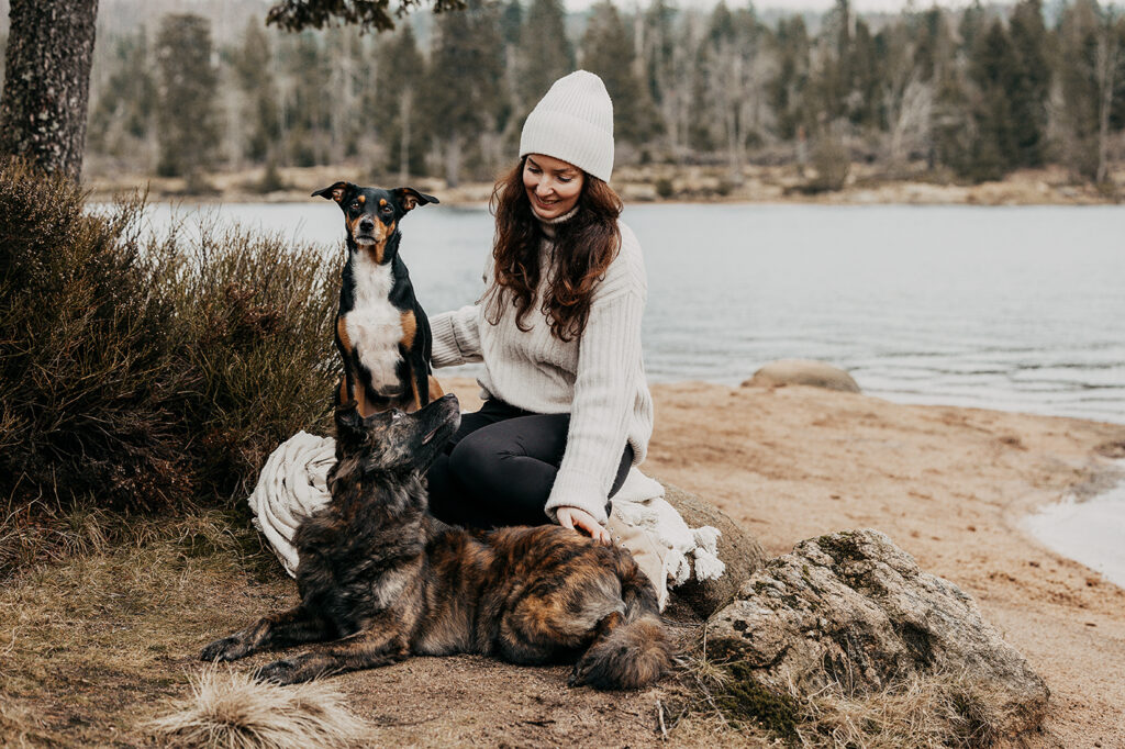 Dunkelhaarige Frau kuschelt liebevoll mit ihren beiden Tierschutzhunden am Oderteich im Harz. Ein Hund bewacht sie aufmerksam, während der andere vertrauensvoll zu ihr aufblickt - eine herzliche Szene voller Vertrauen und Verbundenheit.