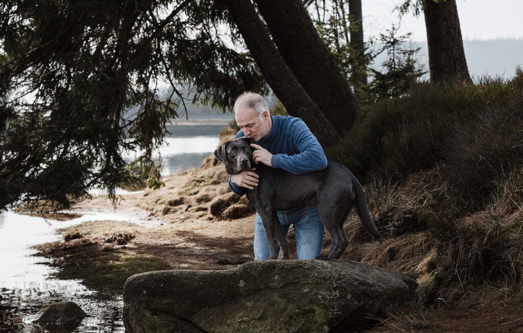 Mann kuschelt liebevoll mit seiner grauen Labrador Hündin am Oderteich im Harz, während sie erwartungsvoll in die Ferne schaut und die Kuscheleinheit genießt