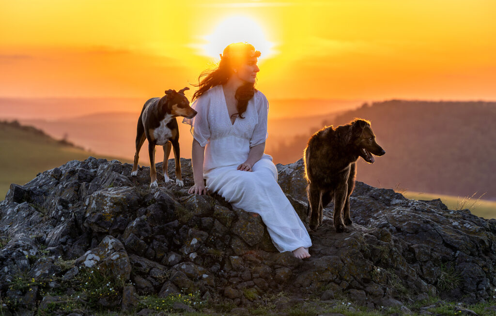 Attraktive braunhaarige Frau in weißem Bohokleid und Trockenblumenhaarkranz, umgeben von ihren treuen Tierschutzhunden, genießt die Himmelsröte und den Sonnenuntergang auf einem Felsen an den Helfensteinen in Dörnberg.