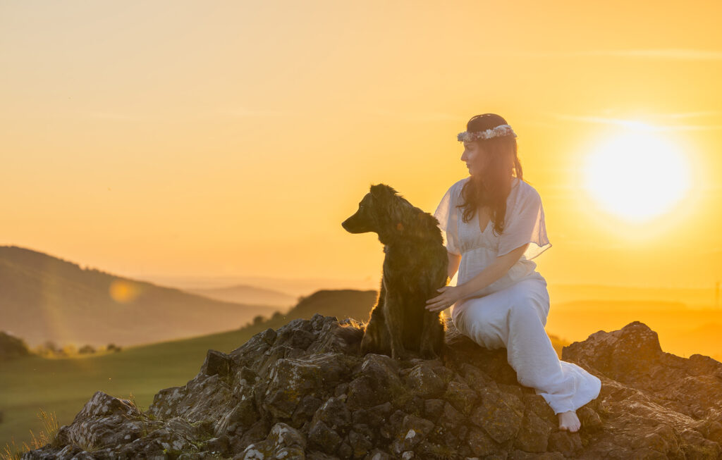 Anmutige braunhaarige Frau in weißem Bohokleid und Trockenblumenhaarkranz, begleitet von ihrem treuen Tierschutzhunden, entspannt sich bei der Himmelsröte und dem Sonnenuntergang auf einem Felsen an den Helfensteinen in Dörnberg.