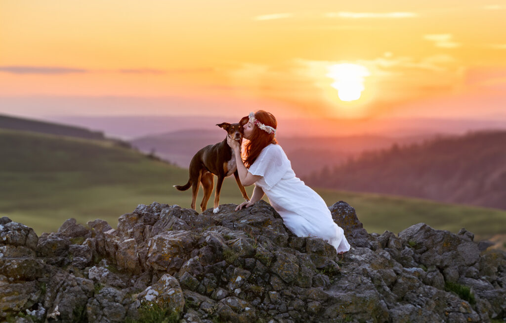 Braunhaarige Frau in weißem Bohokleid und Trockenblumenhaarkranz küsst innig ihre Tierschutzhündin, die den Kuss genießt, auf einem Felsen bei den Helfensteinen in Dörnberg.